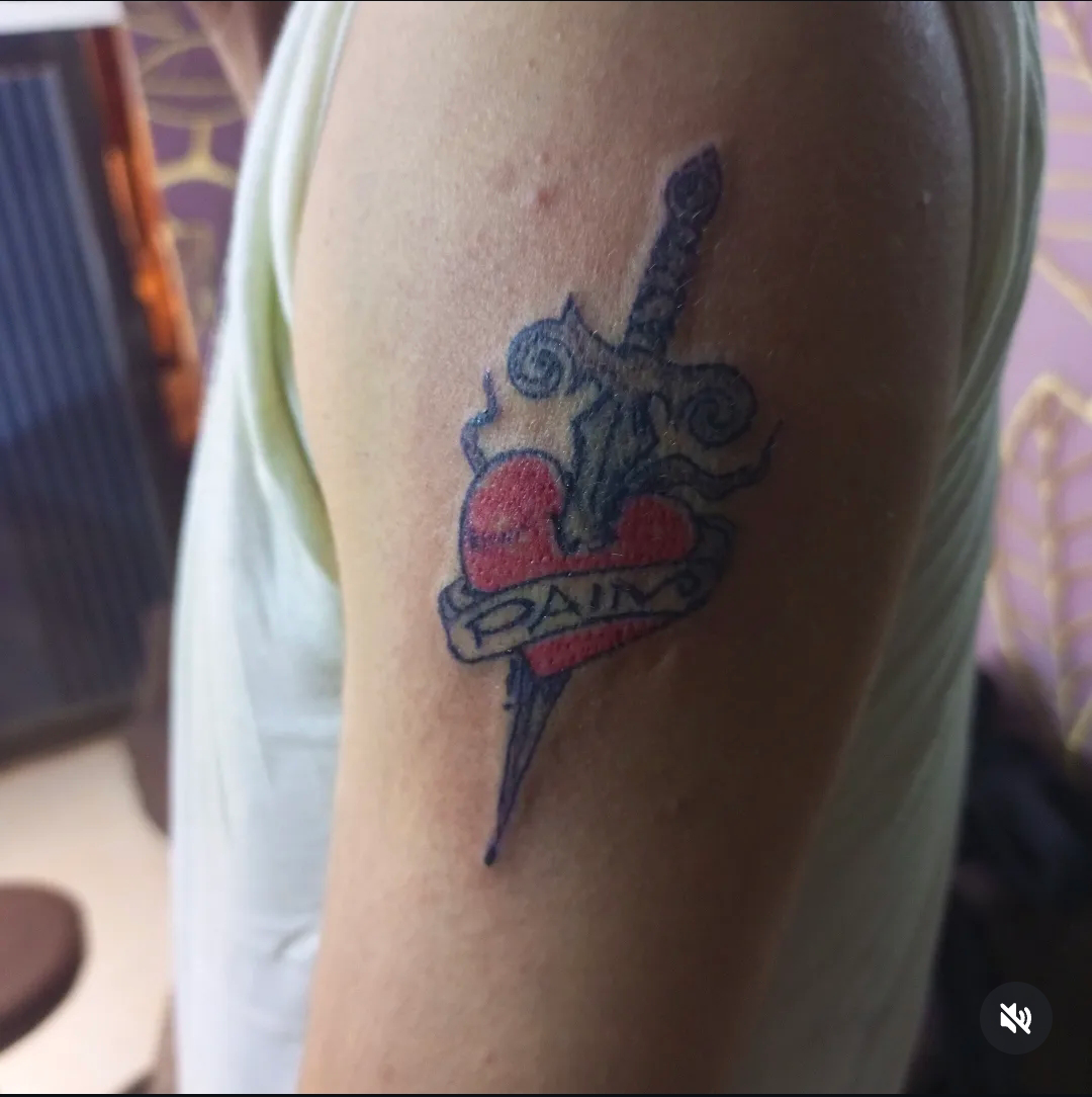 Traditional colour dagger and heart tattoo reading 'Pain' on upper arm, Devil Art Studio Lahore